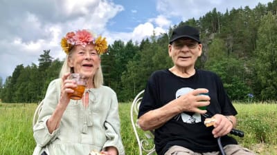 Kaksi vanhusta istuu vierekkäin ulkona, nauttivat juomaa, naisella kukkaseppele päässä.