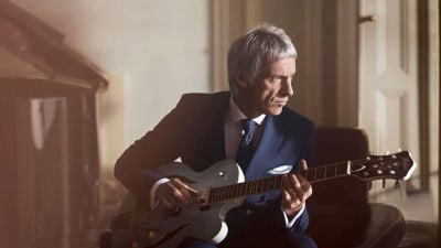 Harmaatukkainen Paul Weller istuu kitara kädessä ja kartselee sivuun.