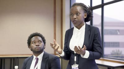 Juristin mustavalkoiseen asuun pukeutunut nuori nainen puhuu käsillään elehtien, vieressä häntä katsoo nuori mies. Kuva dokumentista Afrikan mestariväittelijät.