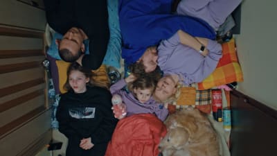 En familj och en hund ligger alla tätt intill varandra på golvet i en skrubb. 
