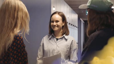 Nainen seisoo toimiston käytävällä ja katsoo hymyillen kahta kameraan selin päin seisovaa kollegaansa.