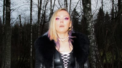 Vaaleahiuksinen ja vahvoihin meikkeihin sonnustautunut nainen seisoo syksyisessä koivumetsässä ja katsoo intensiivisesti kohti kameraa.
