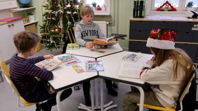 Tre barn sitter runt ett bord och läser böcker.