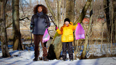 Yle Galaxin Saku ja Lumi hymyilevät keväisessä metsässä kädessään roskapussit ja roskapihdit.