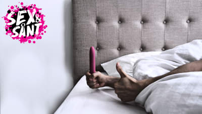 en man som har handen framme vid kanten av sängen under täcket med en dildo i ena handen och med andra handen visar han tummen upp