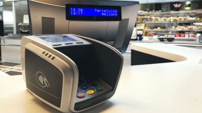 Etualalla yleinen maksupääte, jossa lähimaksuominaisuus. Takana näkyy kassan näyttöruutu ja ruokatiskiä