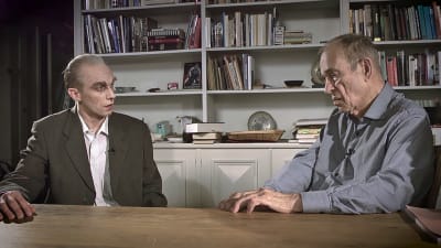 Christoffer Strandberg utklädd till Jörn Donner. Han är sminkad för att se gammal ut samt har stora ögonbryn. Han har också på sig en säckig kavaj och glasögon. Han sitter tillsammans med Jörn i Jörns vardagsrum. I bakgrunden syns böcker. 