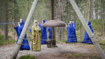 Metsässä sinisiin kaapuihin pukeutuneita naisia, soittavat sinisiä huiluja, keskellä ison kirkonkellon äärellä vihreäkaapuinen nainen naamio kasvoilla. Soivan Metsän Ilmattaria.