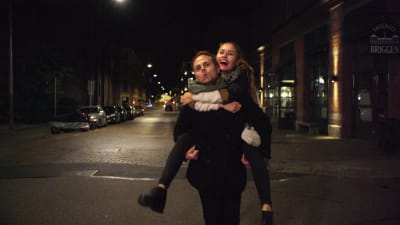 Pojkvännen bär Flickvännen på sina axlar på övergicen storstadsgata på natten.