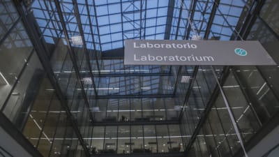 En skylt där det står "laboratorie". I bakgrunden ser man en byggnad med stora fönster.