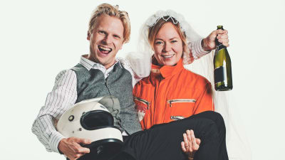 Joppe Dikert iklädd formulaoverall med sin man Oskar Pöysti i famnen. Han har en formel1- hjälm i famnen och en champagneflaska i handen. De ser ivriga och glada ut. 