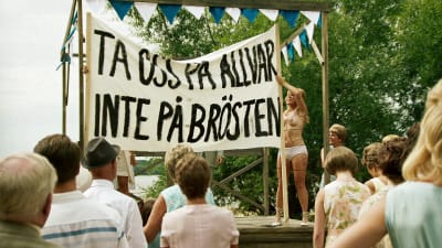 Lottie (Maja Rung) demonstrerar med banderoll: "Ta oss på allvar, inte på brösten"