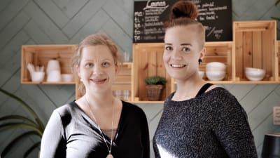 Två medelålders kvinnor i ett kafè ler och tittar in i kameran 