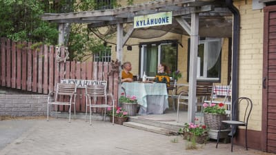 Man och kvinna sitter och äter vid ett bord på en terass framför ett ljusgult hus. Ovanför terassen en skylt med texten "Kylähuone". 