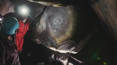Två personer inuti en mörk grotta lyser upp en solliknande grottmålning med sina pannlampor.