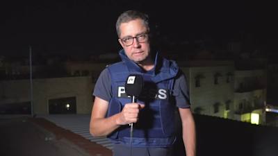 En reporter med Yle-mikrofon står med en skottsäker väst på ett tak. Det är kväll.