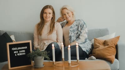 Två kvinnor sitter i en soffa och ler. De är båda gravida. 