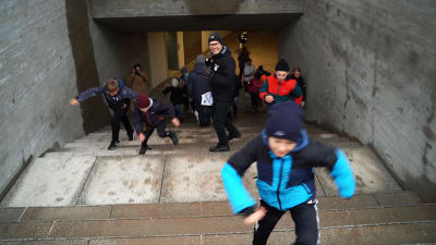Glad barn springer upp för en tunnel och en redaktör med en mikrofon i handen står mitt i bild och ser glad ut.