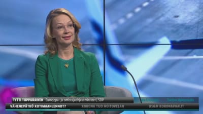 Europa- och ägarstyrningsminister Tytti Tuppurainen klädd i grönt ser in i kamera och ler. Hon sitter i Yles tv-studio där bakgrunden är blå och abstrakt.