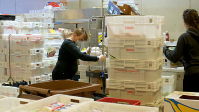 Kvinna omgiven av vita plastbackar i matutdelningshall i Kvarnbäcken Helsingfors