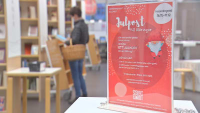 En röd skylt på ett bord gör reklam för kampanjen Julpost till åldringar. I bakgrunden syns en person med korg på armen som tittar på julkort i ett bibliotek.