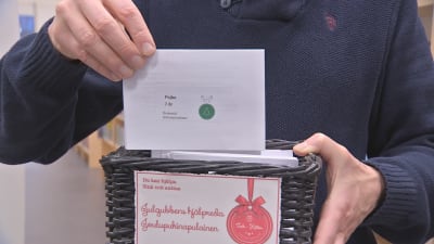 En person med blå tröja drar upp ett kort ur en liten låda med julklappsönskemål. På lappen står att en 7-årig pojke önskar sig små plastsoldater.