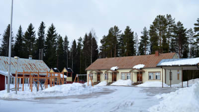 Skolgård täckt i snö. På bilden ett gammalt trähus som är beigemålat, framför liten lekpark med klätterställning och gungor.