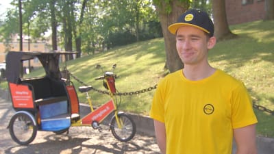 En man med gul skjorta och keps står vid sin cykeltaxi, riksha, vid en park en varm sommardag.
