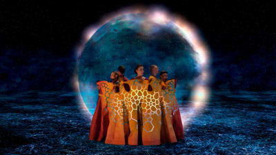 Tanssijoita oransseissa kaavuissa, taustalla sinisenä hehkuva planeettamainen taivaankappale.
