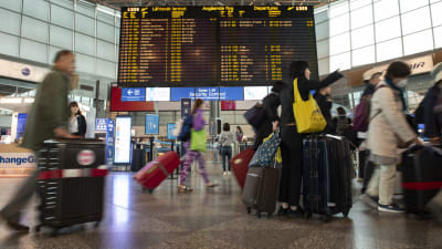 Resenärer med kappsäckar går på Helsingfors-Vanda flygplats. I mitten syns avgående flyg på en tavla.