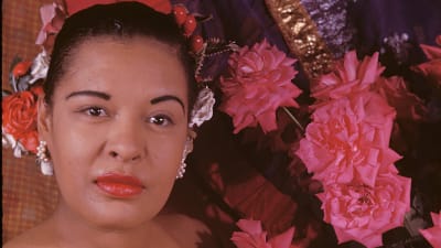 Laulaja Billie Holiday lähikuvassa, taustalla kukkia.
