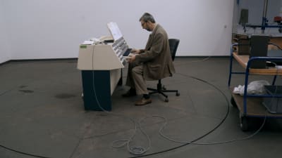 Mies istuu tyhjässä huoneessa ja soittaa oudon näköistä syntetisaattoria, kuva dokumenttielokuvasta Subharchord.