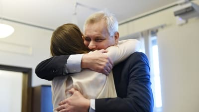Antti Rinne, en äldre man med grått hår i kostym omfamnar Sanna Marin. Kvinnan har en ljus tröja och brunt hår.