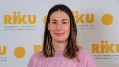 en kvinna i brunt halvlångt hår och rosa tröja står framför en vägg med ordet "riku" på.