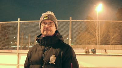 Porträttfoto av man som står framför en ishockeyrink. Mannen har mössa, glasögon och dunjacka. Det är vinter och mörkt ute.