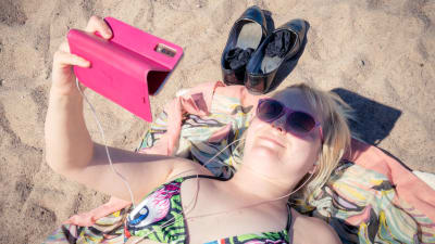 Kvinna på stranden. Hon ligger och tittar på video via sin mobiltelefon.