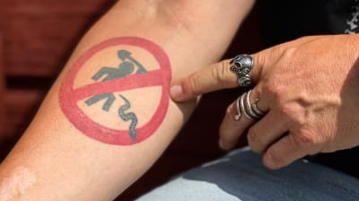 närbild på en arm med en tatuering med ett förbudsmärke över en streckgubbe som dödar en orm.