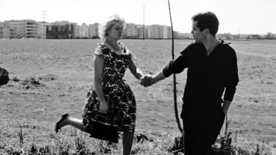 Kukkamekkoinen vaalea nainen ja mustapaitainen mies rientävät käsi kädessä aukealla ruohokentällä, kaukana taustalla kaupungin kerrostaloja. Kuva Pasolinin elokuvasta Pummi.