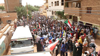 Människor viftar med Sudans flagga och protesterar. En man står på en buss och fotograferar demonstrationen.
