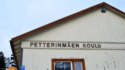 Närbild av en gammal träbyggnad, beige färg, det står Petterinmäen koulu på väggen.