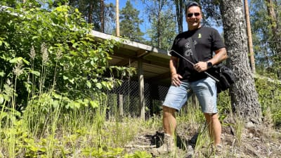 En man i t-skjorta och shorts poserar med en ormkrok i handen framför ett litet hus.
