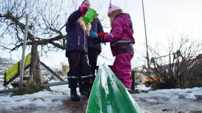 Barn samt en barnskötare leker med vatten i en grön vattenränna i slaskigt vinterväder.