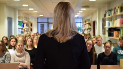 Sandra bergqvist med ryggen till kameran står framför en gymnasieklass och talar.