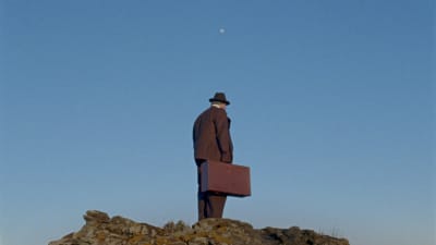 Hattupäinen mies seisoo vanhanaikainen matkalaukku kädessään kalliolla ja katsoo jonnekin; yllä sinisellä taivaalla on kuu.