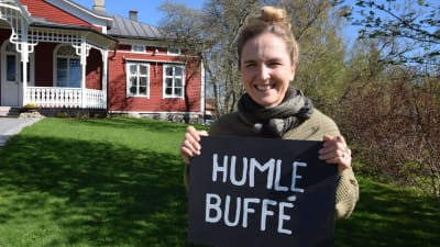 Framför ett rött hus står en leende kvinna med en skylt "Humle Buffet"