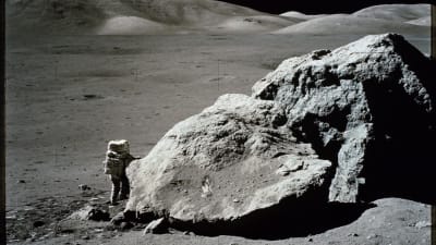 Astronautti seisoo Kuussa suuren kiven vieressä.