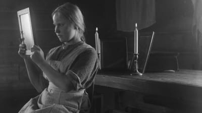 Svartvit bild från början av 1900-talet på en flicka som sitter vid ett bord med två levande ljus och trollbunden tittar in i en spegel som hon håller i med bägge händerna.