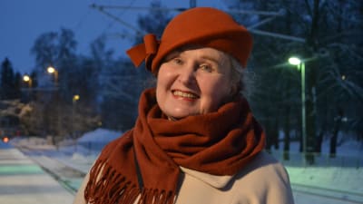 Porträttfoto av dam tagen utomhus vintertid. Damen har på sig en rostbrun hatt och en rostbrun halsduk till en beige kappa. Hon ler.