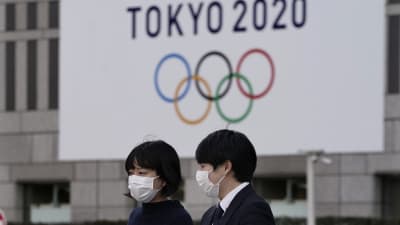 två personer står med ansiktsmasker framför en OS-skylt.