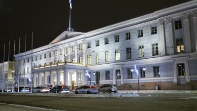 Helsingfors stadshus. Det är en mörk vinterkväll. På gatan utanför stadshuset står bilar på rad. På gatan ligger snö.